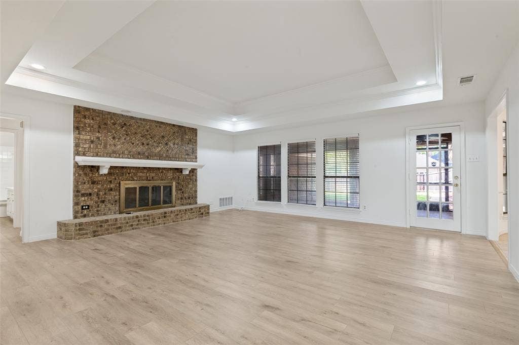 Spacious living area featuring a brick fireplace with a white mantel, light wood-finish flooring, a tray ceiling, and multiple windows with blinds