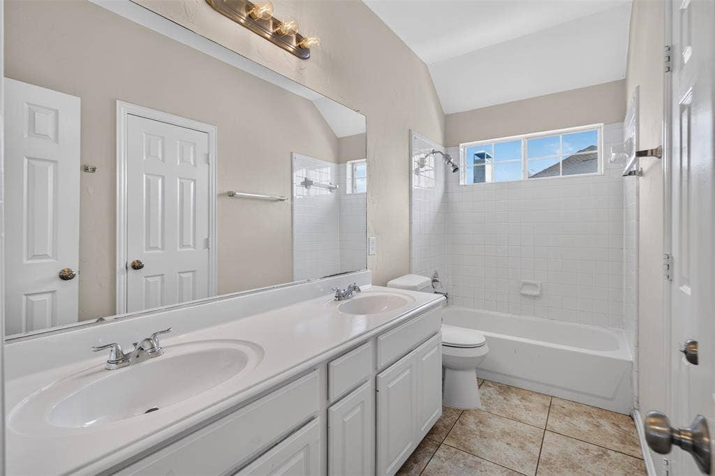 7112 Chandler Drive, Plano, TX 75024 Double vanity with white cabinetry and integrated sinks, large wall-mounted mirror, ceramic tile flooring, bathtub with shower, and an arched ceiling