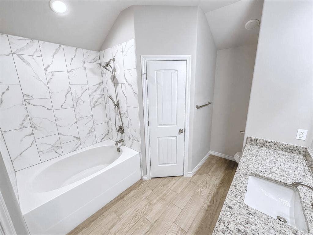 1210 Campbelton Drive, Temple, TX 76504 Bathroom featuring a built-in tub with a shower head, white marble-style wall tiling, a light wood-finish floor, a white vanity with an undermount sink, and a speckled countertop