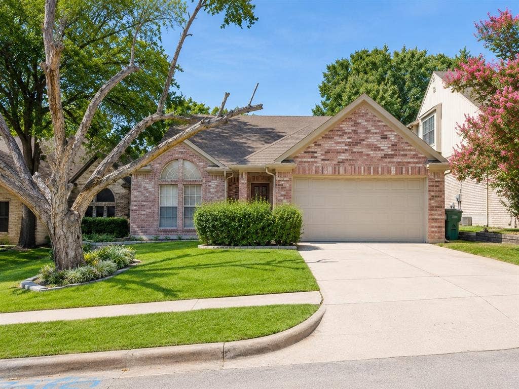 1106 Cain Court, Cedar Hill, TX 75104 Brick facade residence featuring a two-car garage, concrete driveway, arched windows, and established landscaping with mature trees and shrubs