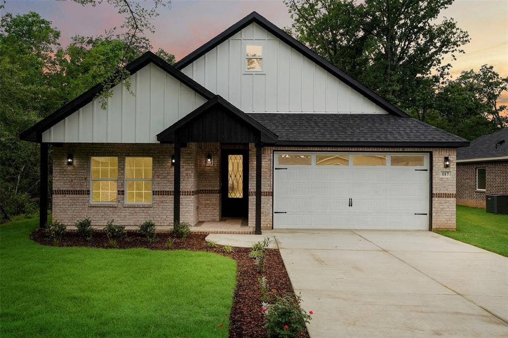 117 Parkside Drive, Trinidad, TX 75163 Exterior featuring a brick facade, white board and batten siding, dark shingle roof, attached garage with decorative hardware, and a landscaped front yard with a concrete driveway