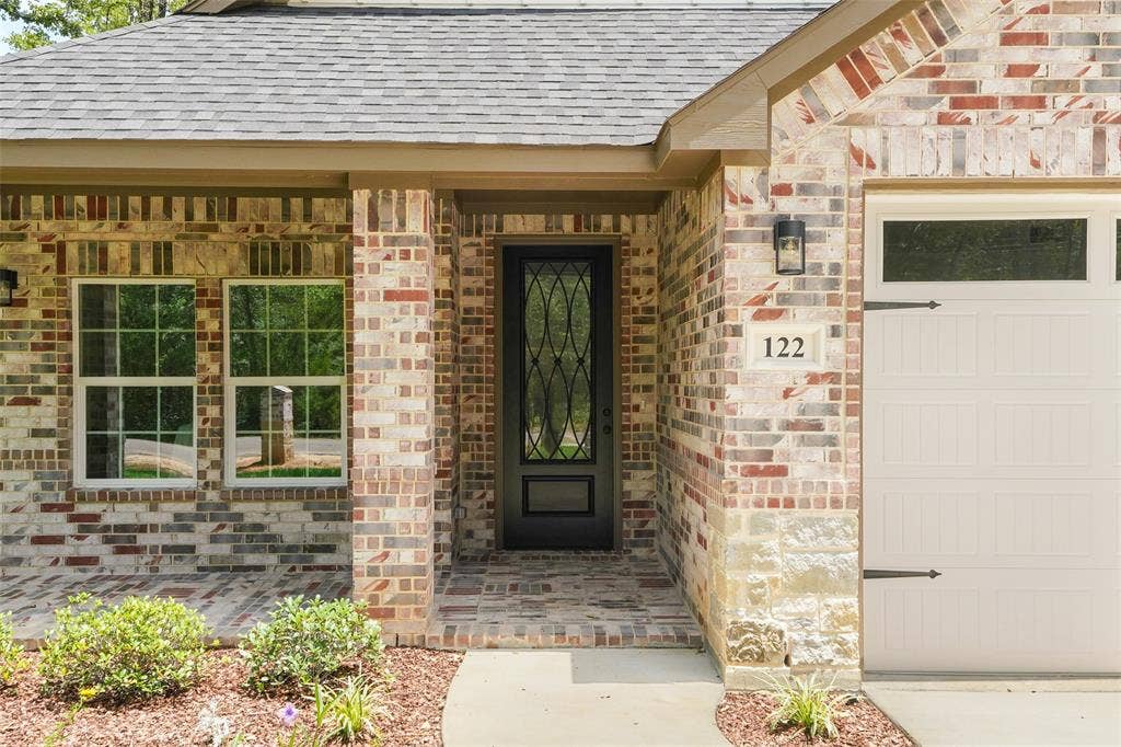 122 E Beachwood Loop, Trinidad, TX 75163 Brick exterior featuring a covered porch with a patterned brick floor, a dark-paneled entry door with decorative glass, two double-hung windows, and an attached garage with a window panel and decorative hardware