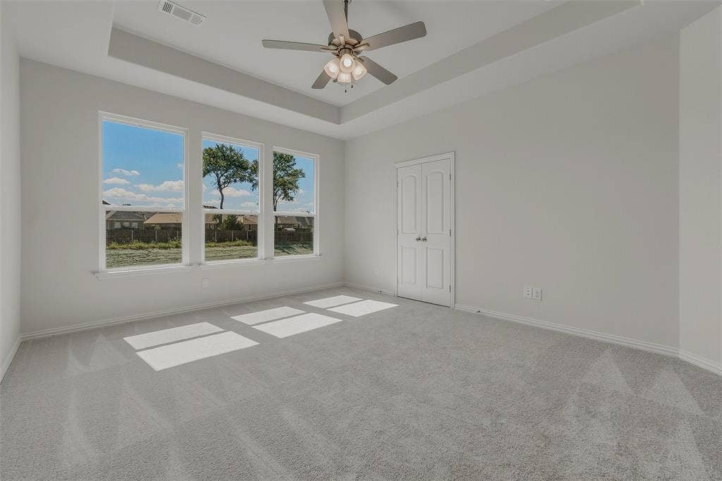 Tray ceiling with a ceiling fan, three expansive windows, light gray carpeting, and a double-door closet