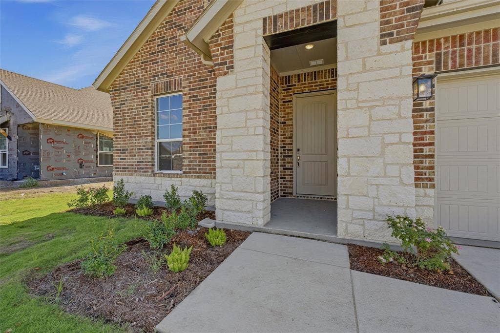 Brick and stone exterior with a covered entry, concrete walkway, attached garage, and landscaped front yard