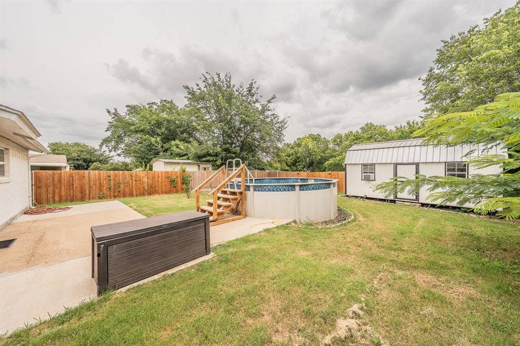 5612 Macgregor Drive, Haltom City, TX 76148 Backyard featuring an above-ground pool with a wooden deck and stairs, a concrete patio, a wooden privacy fence, and a white storage shed with a metal roof
