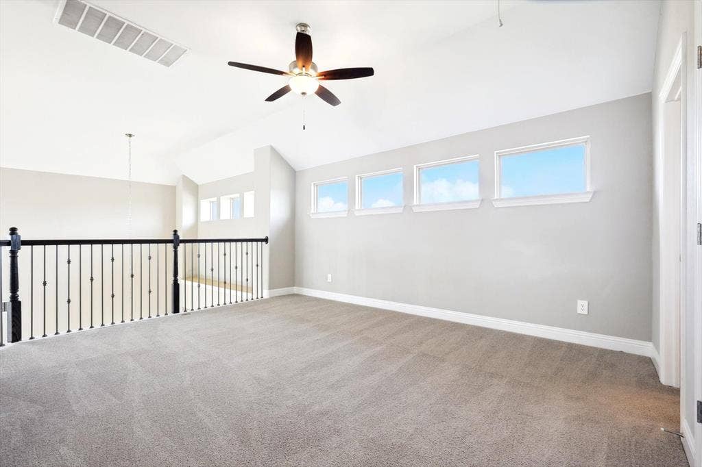 Spacious loft area featuring vaulted ceilings with a ceiling fan, three horizontal windows with white trim, light gray wall paint, and gray carpeted flooring