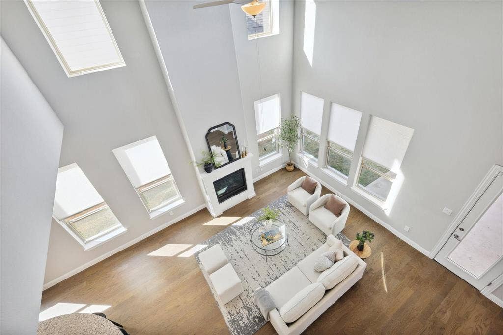 Two-story living area featuring high ceilings, light wood-finish flooring, multiple windows with blinds, a white fireplace with mantle, and a ceiling fan