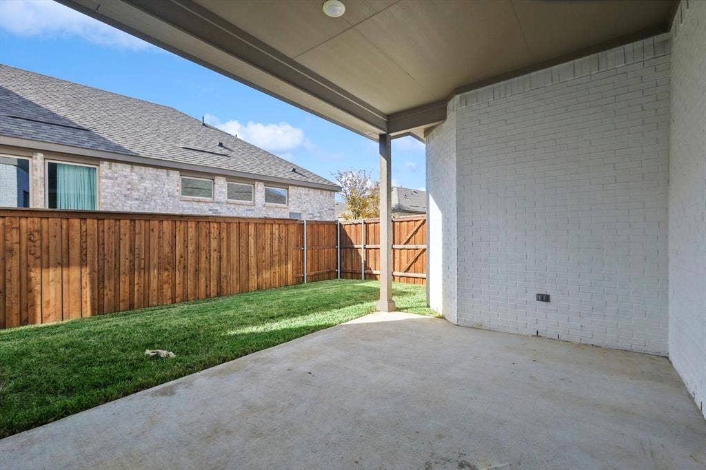 Covered patio with a concrete surface, featuring white brick exterior walls and a fenced yard with natural wood-tone fencing and grass