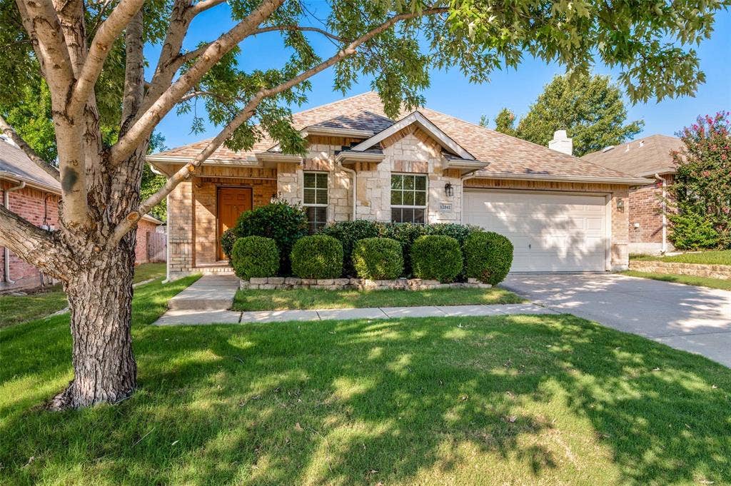 12841 Serenity Drive, Frisco, TX 75035 Stone and brick facade home featuring a two-car garage, mature front yard tree, concrete driveway, and established front landscaping