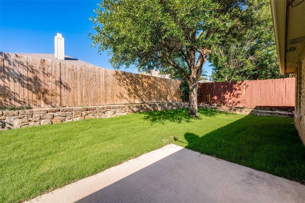 12841 Serenity Drive, Frisco, TX 75035 Private backyard featuring a concrete patio, a mature tree providing shade, and a stone retaining wall