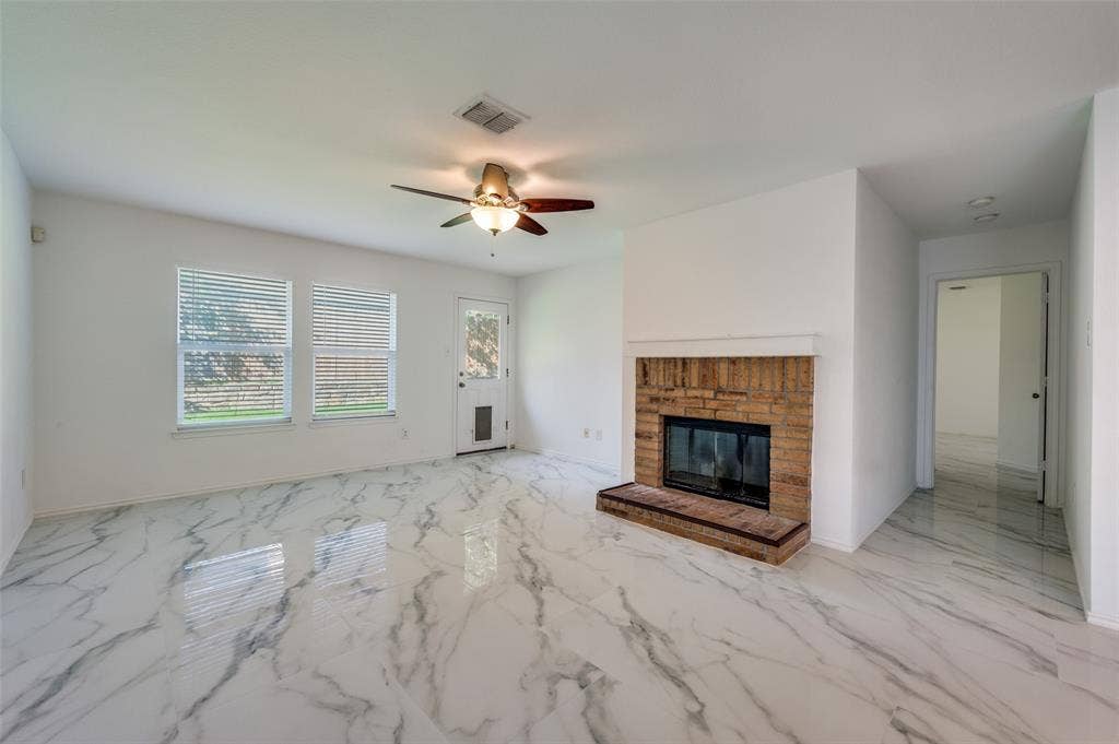 12841 Serenity Drive, Frisco, TX 75035 Spacious living area featuring polished marble-look tile flooring, a brick fireplace with hearth, a ceiling fan with integrated lighting, and two windows providing natural light