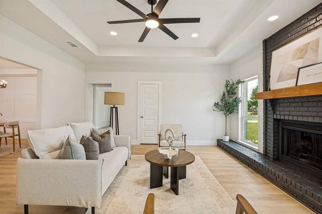 109 Shadow Drive, Red Oak, TX 75154 Living area featuring a black brick fireplace with a wood mantel, light wood-finish flooring, a tray ceiling with recessed lighting and a ceiling fan, and a large window