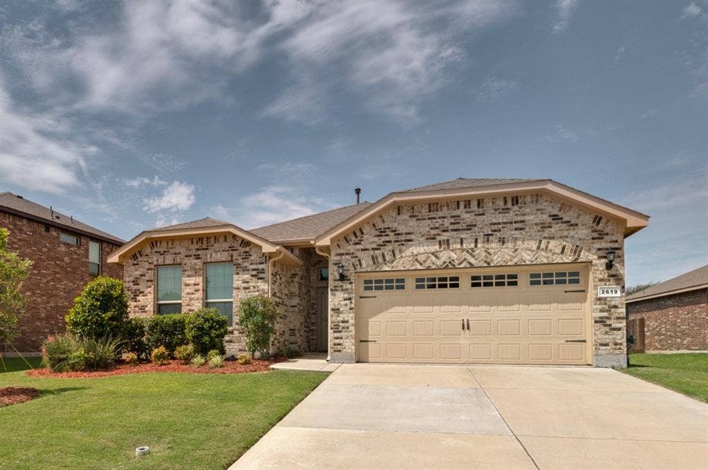 Single-story brick exterior featuring a two-car garage with decorative hardware, a concrete driveway, and a manicured lawn with mature shrubs