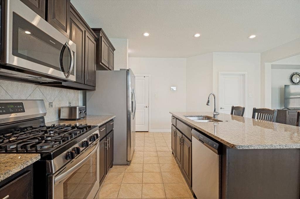 Kitchen featuring dark wood cabinetry, stainless steel appliances, light-toned granite countertops, and tile flooring