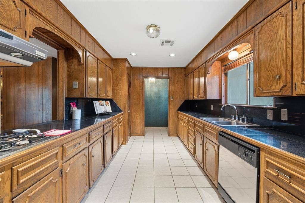 Galley kitchen featuring extensive wood cabinetry, dark countertops, white tile flooring, stainless steel gas range, and a stainless steel dishwasher