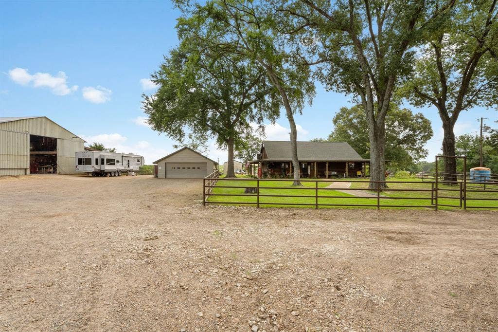 13494 County Road 2919, Eustace, TX 75124 Expansive gravel driveway providing extensive parking, a large metal barn, a detached garage with an overhead door, and a ranch-style home featuring a covered front porch, all surrounded by mature trees and a fenced perimeter