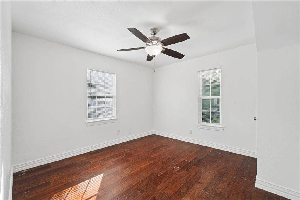 Wood-finish flooring with white baseboards, two windows with white trim, and a ceiling fan with integrated lighting