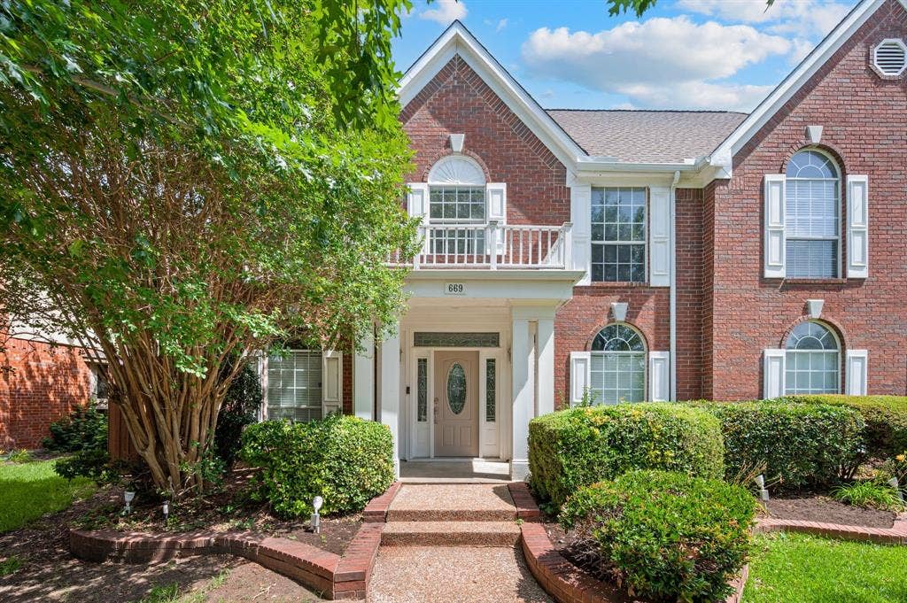 669 Water Oak Drive, Plano, TX 75025 Brick facade residence featuring a covered entry, arched windows with shutters, an upper-level balcony, and mature landscaping