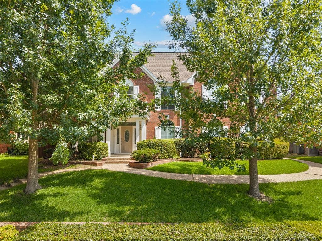 669 Water Oak Drive, Plano, TX 75025 Traditional brick facade featuring white columns and shutters, an arched entry door, a multi-pane window, mature trees, and a curved paved walkway