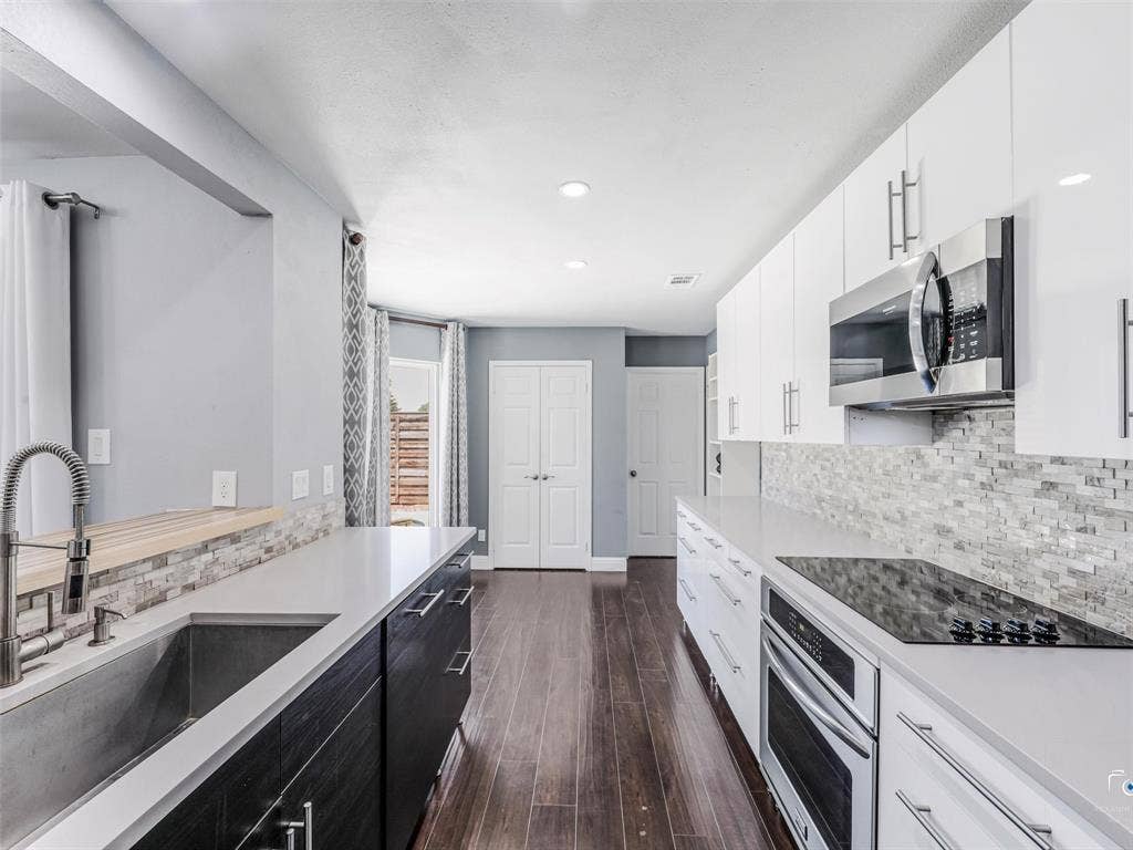 6701 Vero Drive, Plano, TX 75023 Modern kitchen featuring wood-finish flooring, white flat-panel upper cabinetry, dark flat-panel lower cabinetry, light-toned countertops, and a full-height tile backsplash. The pantry is at the left double doors and the laundry room is beyond the door to the right. Note teh large sink!