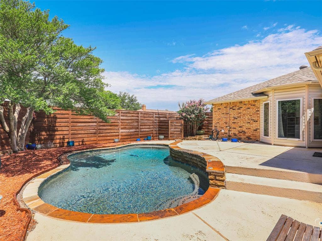 6701 Vero Drive, Plano, TX 75023 Curved swimming pool with stone coping, surrounded by a concrete patio and flower beds.