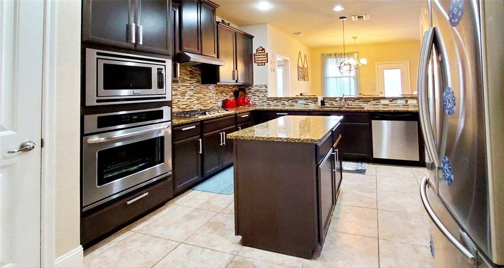 104 Rustic Oak Lane, McKinney, TX 75072 Kitchen featuring dark wood cabinetry, granite countertops, an integrated stainless steel oven and microwave combination, a mosaic tile backsplash, and a central island with additional storage