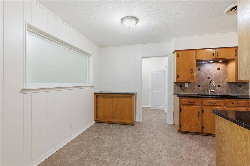 Kitchen featuring tile flooring, wood cabinetry, dark countertops, and a tile backsplash with accent inserts