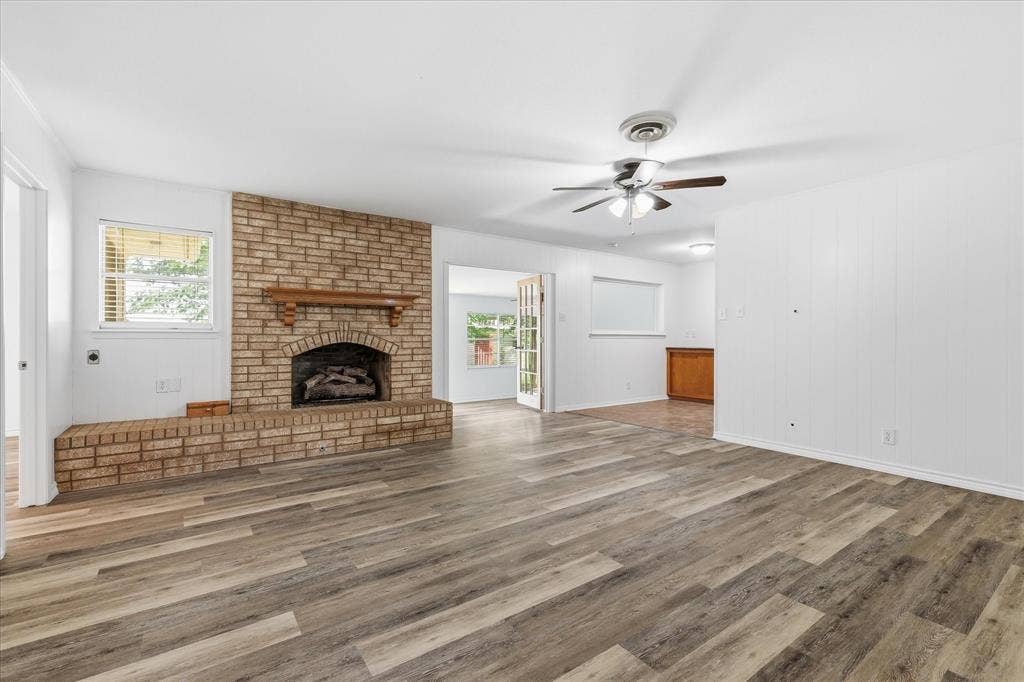 Spacious room featuring wood-finish flooring, a brick fireplace with a wood mantel, a ceiling fan, and white walls
