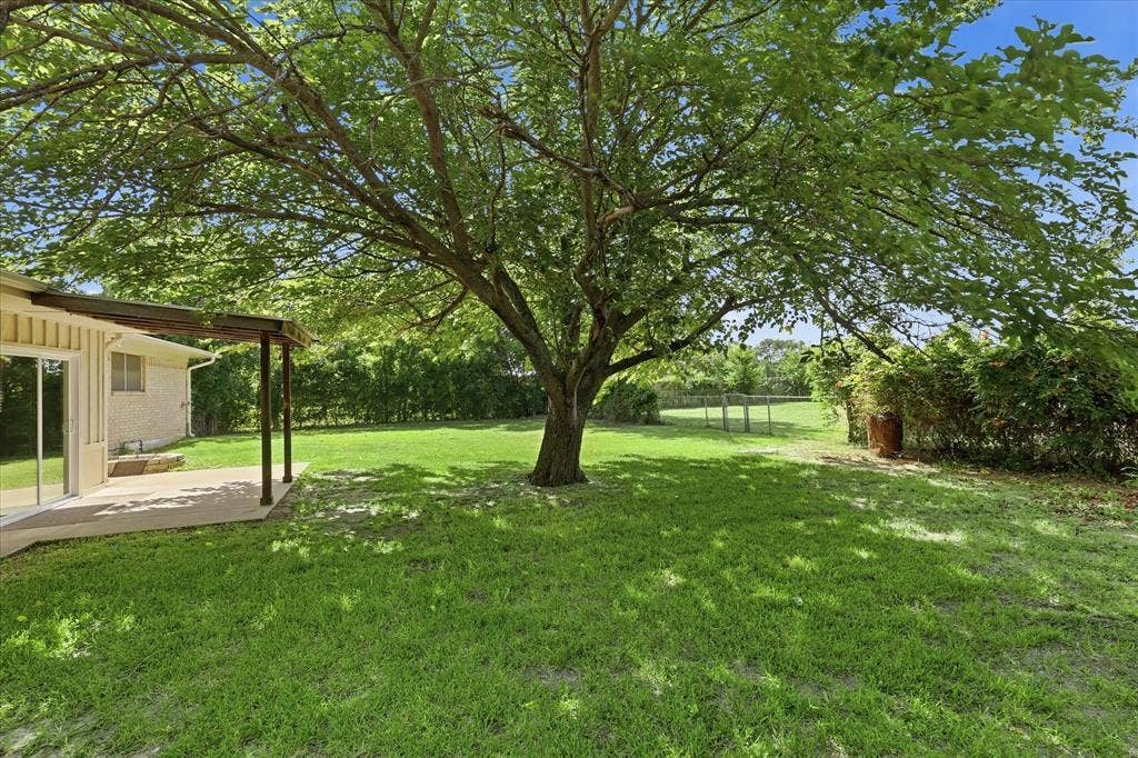 Expansive backyard featuring a mature tree providing natural shade, a large green lawn, a covered patio, and a sliding glass door entry to the residence