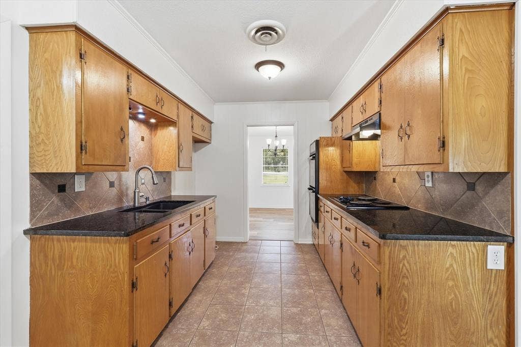 Galley kitchen featuring wood cabinetry, black countertops, a stainless steel faucet, and a patterned tile backsplash