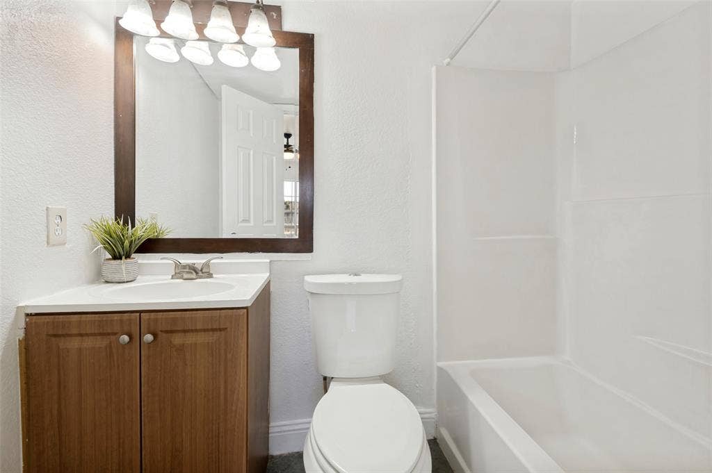 403 N Britain Road, #407B, Irving, TX 75061 Bathroom featuring a wood-finish vanity with an integrated sink and chrome faucet, a framed mirror with a four-bulb light fixture, a toilet, and a white bathtub with a shower surround