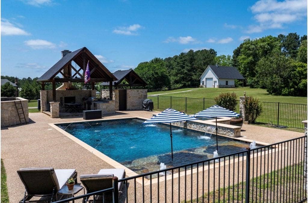 106 Timber Hill, Athens, TX 75751 Expansive outdoor living area featuring a custom pool with water features, a built-in outdoor kitchen with stone detailing and a wood-beamed pavilion, a gravel patio, and a black wrought-iron fence