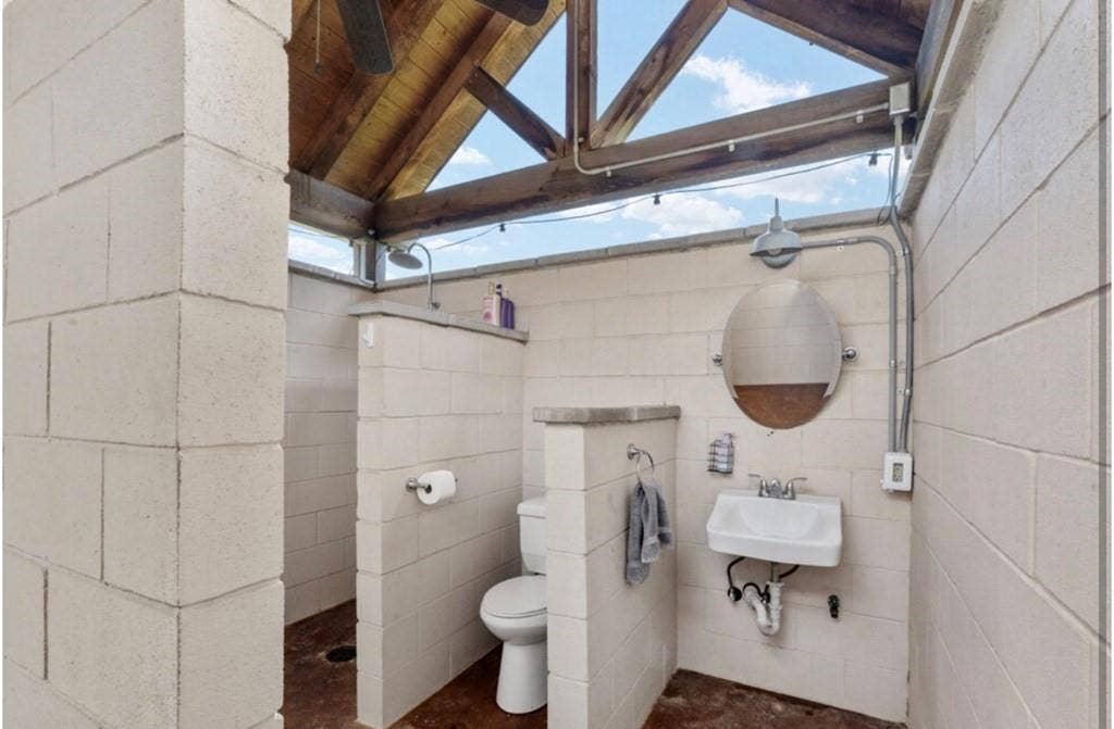 106 Timber Hill, Athens, TX 75751 Outdoor bathroom featuring concrete block walls, exposed wood beam ceiling, open-air showering area, and a wall-mounted sink with an oval mirror