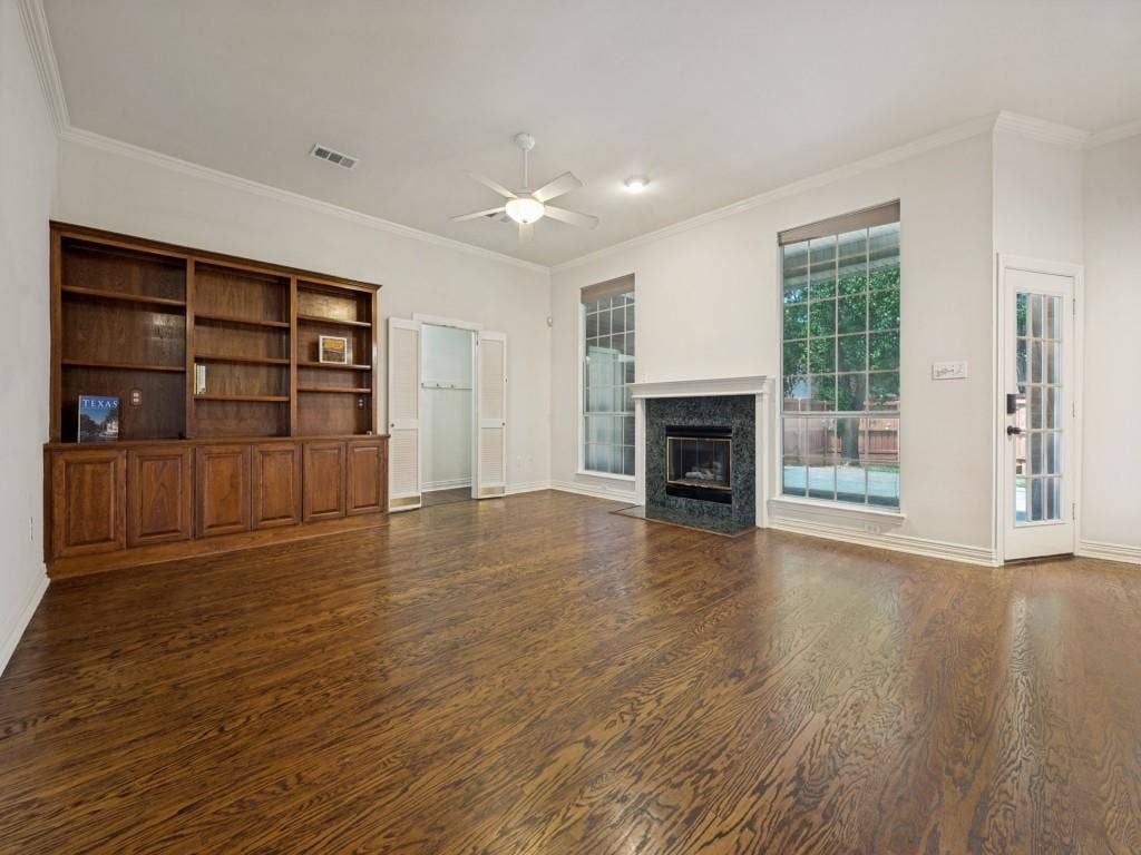 9703 Raven Lane, Irving, TX 75063 Spacious room featuring wood-finish flooring, a built-in wood shelving unit, a fireplace with a dark stone surround, two large windows, and a glass-paned door