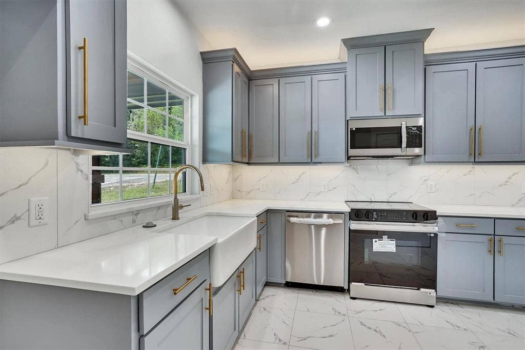 Modern kitchen featuring light gray blue cabinetry, gold-tone hardware, quartz white countertops, a white farmhouse sink, and a stainless steel appliance package