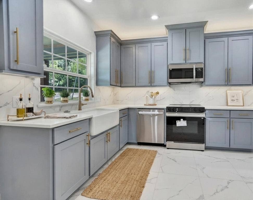 Modern kitchen featuring light gray cabinetry with gold hardware, white countertops, a deep farmhouse sink, stainless steel appliances, and white marble-look tile flooring