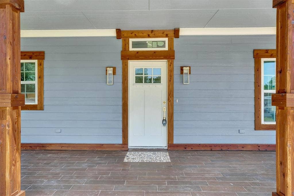 Inviting front porch featuring wood-finish flooring, light gray siding, white entry door with transom window, and natural wood trim accents