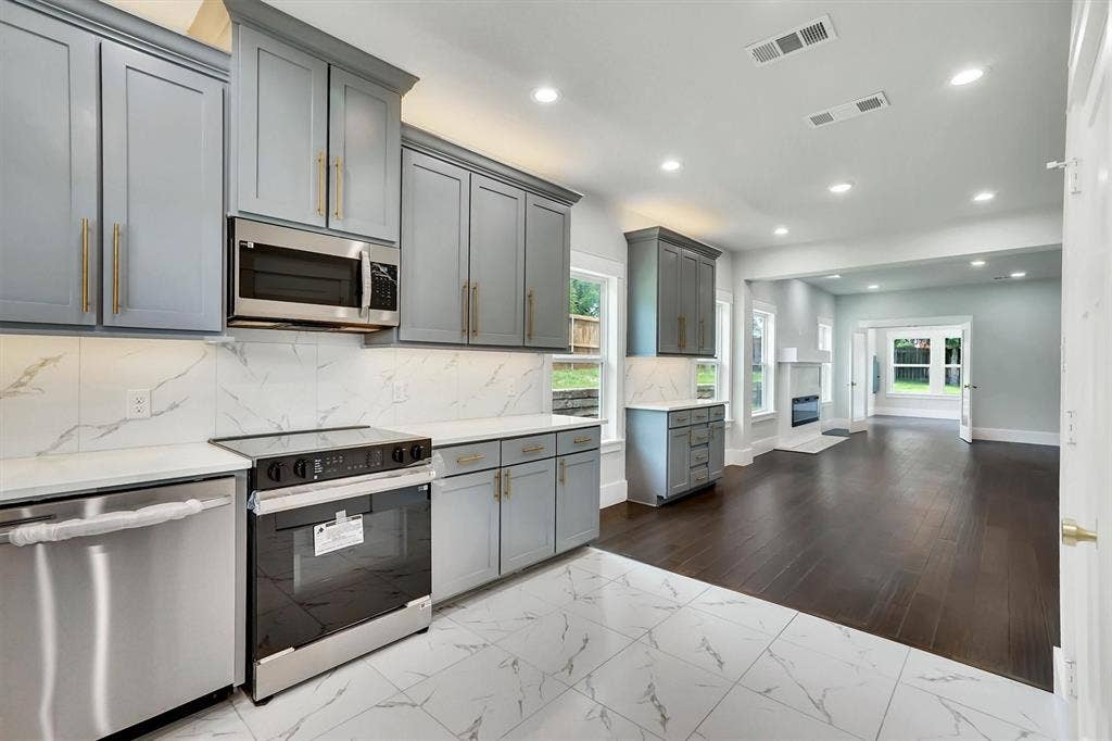 Modern kitchen featuring stainless steel appliances, light gray cabinetry with gold-tone hardware, quartz countertops, and a white tile backsplash