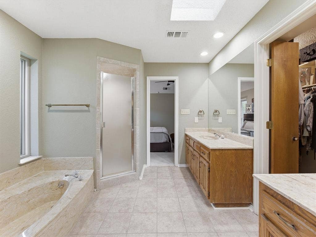3701 Waycross Court, Arlington, TX 76016 Primary Bathroom featuring a skylight, tile flooring, a built-in tub with stone surround, a glass-enclosed shower, and a dual vanity with a light-toned countertop