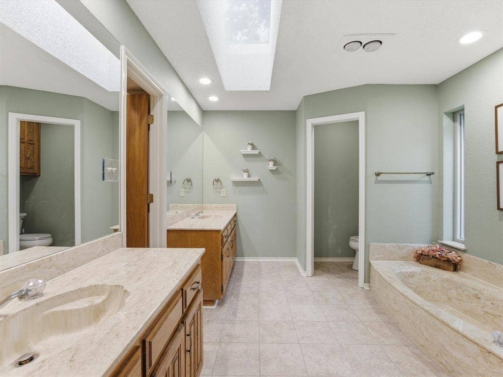 3701 Waycross Court, Arlington, TX 76016 Primary bathroom featuring a large skylight, dual vanity with integrated sinks, wood-finish cabinetry, a built-in tub with stone-finish surround, and tiled flooring
