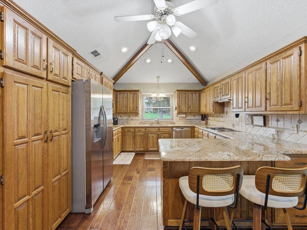 3701 Waycross Court, Arlington, TX 76016 Kitchen featuring a vaulted ceiling with exposed wood beams, extensive wood cabinetry, wood-finish flooring, a stainless steel refrigerator, and stone countertops