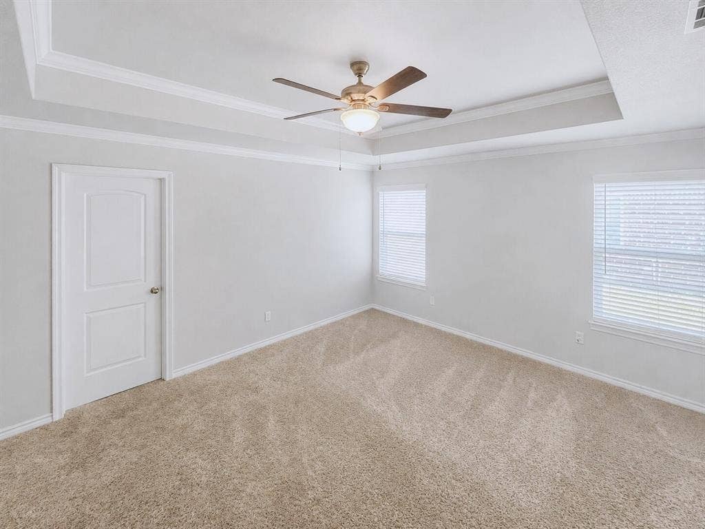 Spacious room featuring tray ceilings with crown molding, a ceiling fan with integrated lighting, two windows with blinds, and light-toned carpeting