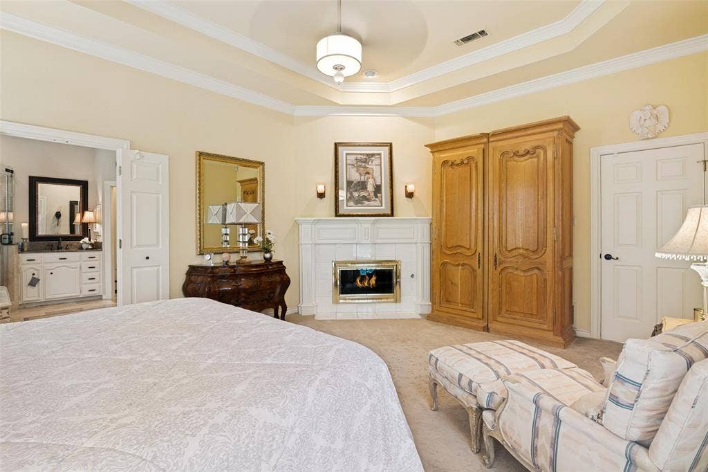 Recessed ceiling with layered crown molding, corner fireplace with white mantel and hearth, built-in wooden armoire, and an entryway to an ensuite bathroom with a double vanity