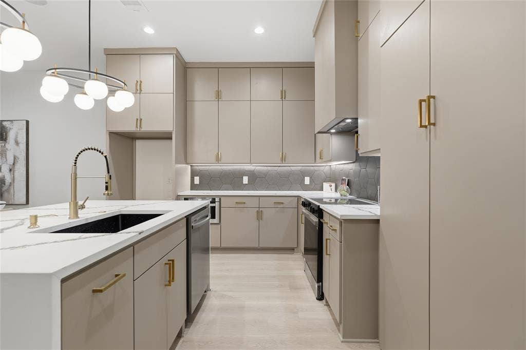 Open-concept kitchen featuring light-toned cabinetry with gold-finish hardware, a large island with a black sink and gold-finish faucet, white stone countertops, and a gray hexagonal tile backsplash