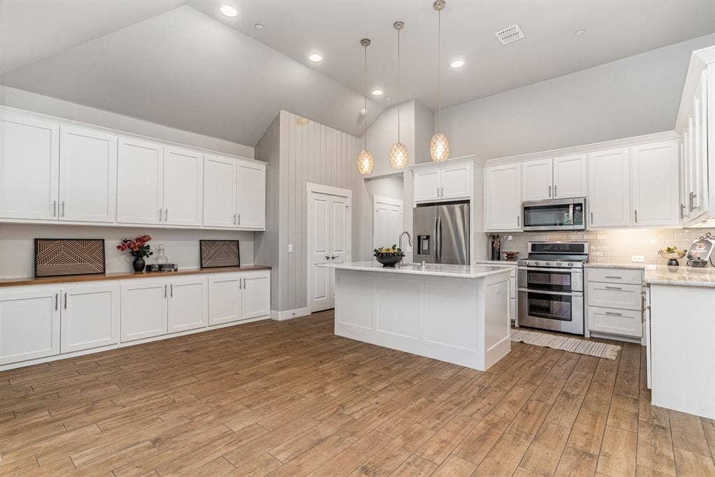 3861 Dalea Drive, Prosper, TX 75078 Open-concept kitchen and living area featuring wood-finish flooring, abundant white cabinetry, a central island with a sink, stainless steel appliances, and vaulted ceilings with recessed lighting