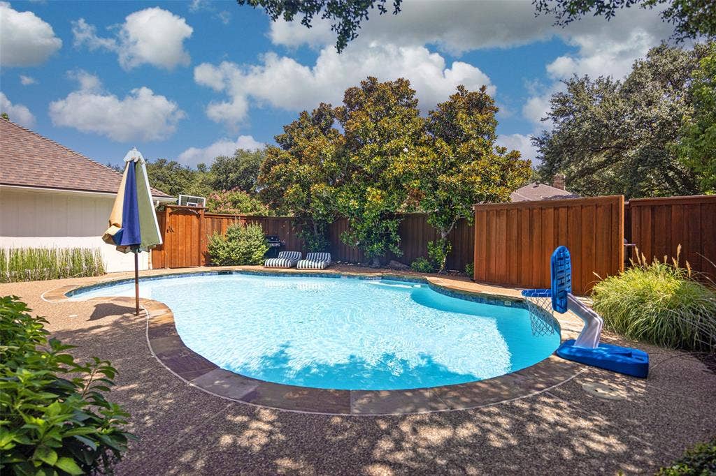 6501 Copper Creek Drive, Dallas, TX 75248 Curved swimming pool with an exposed aggregate deck, bordered by mature trees and privacy fencing