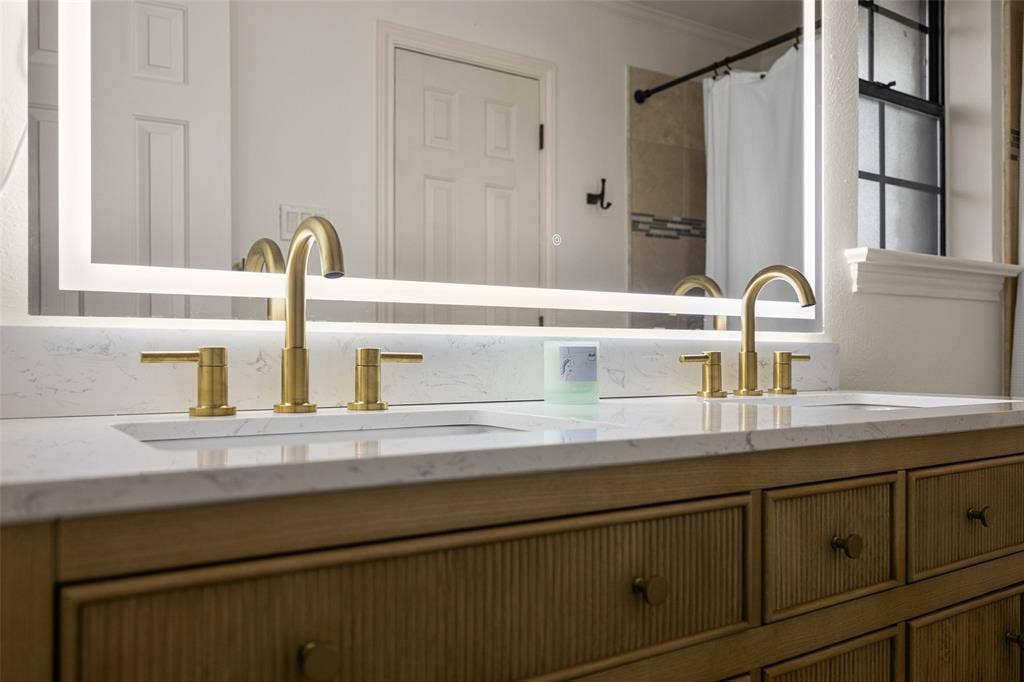 6501 Copper Creek Drive, Dallas, TX 75248 Dual vanity featuring a white countertop with integrated sinks, brushed gold faucets, and a large backlit mirror