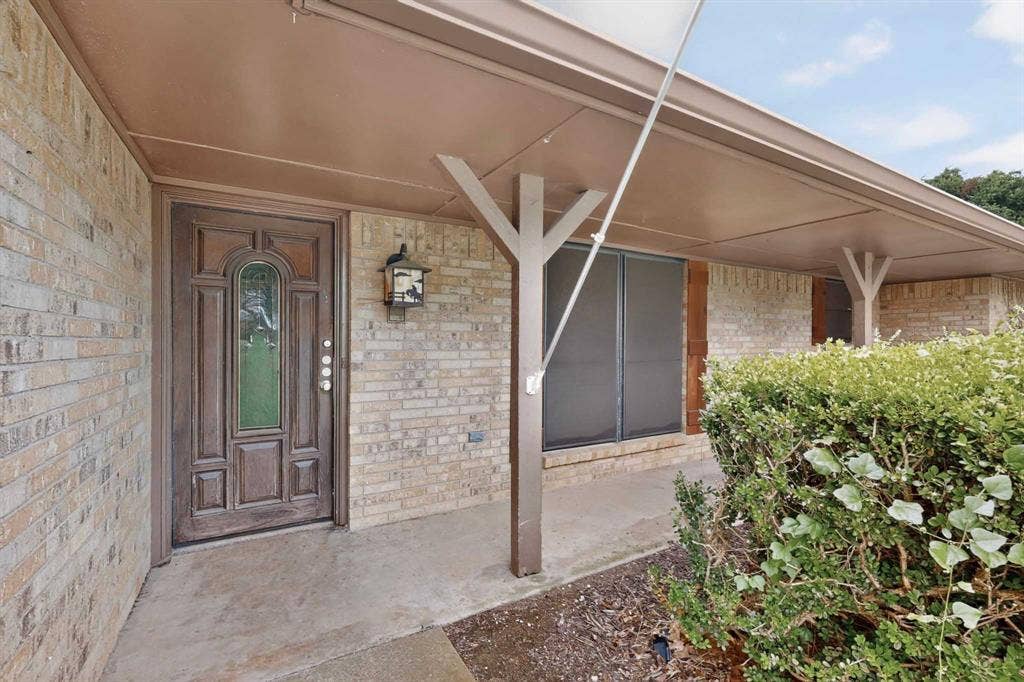 Covered entry with patterned wood front door featuring an arched glass insert, brick exterior, and a built-in exterior light fixture