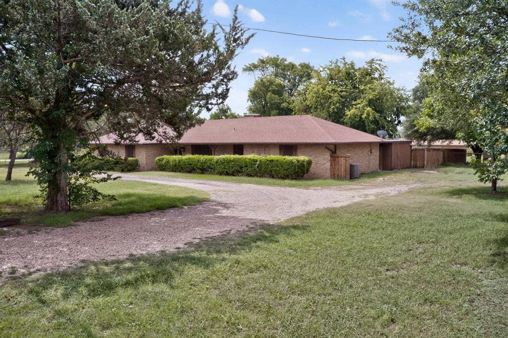 Exterior brick residence with a gravel driveway, mature trees, manicured shrubbery, and a brown shingle roof