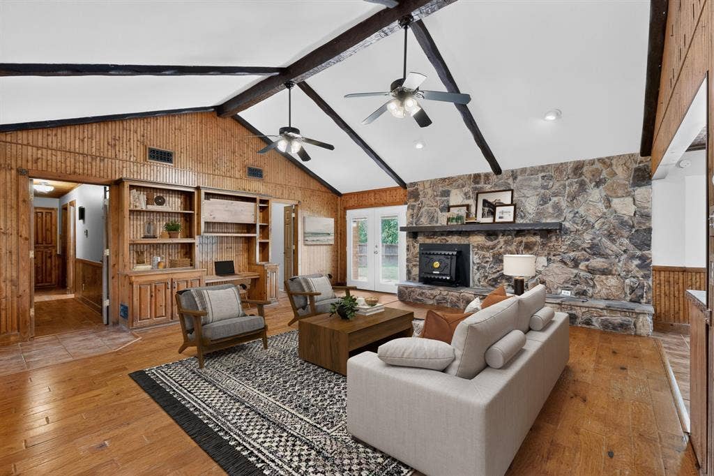 Spacious living area featuring a stone accent wall with a built-in fireplace, vaulted ceilings with exposed beams and ceiling fans, wood-finish flooring, and built-in shelving with cabinetry
